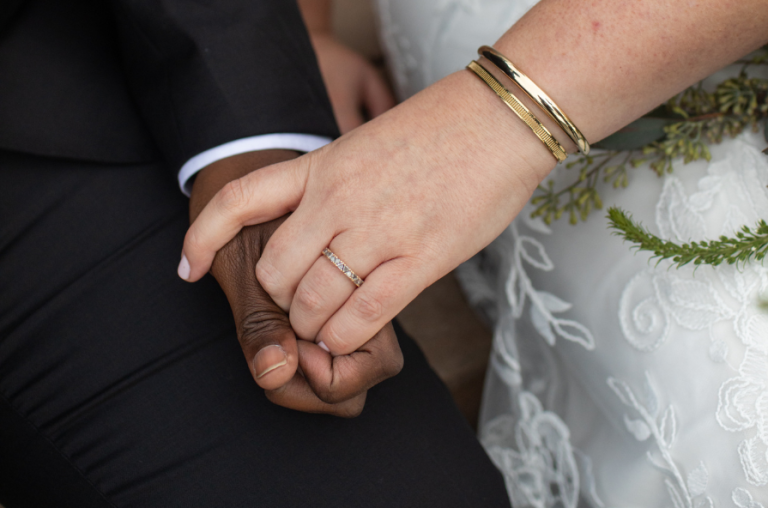 Interracial newlyweds hold hands with their wedding bands.