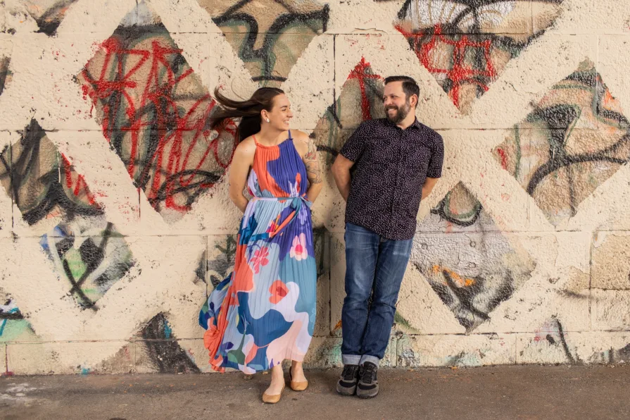 Two engaged folks smiling in Schuylkill River Park in front of a mural.