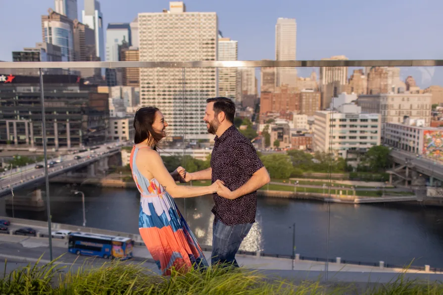 An engaged couple smiling and dancing in front of the Philadelphia skyline at Cira Green's rooftop .