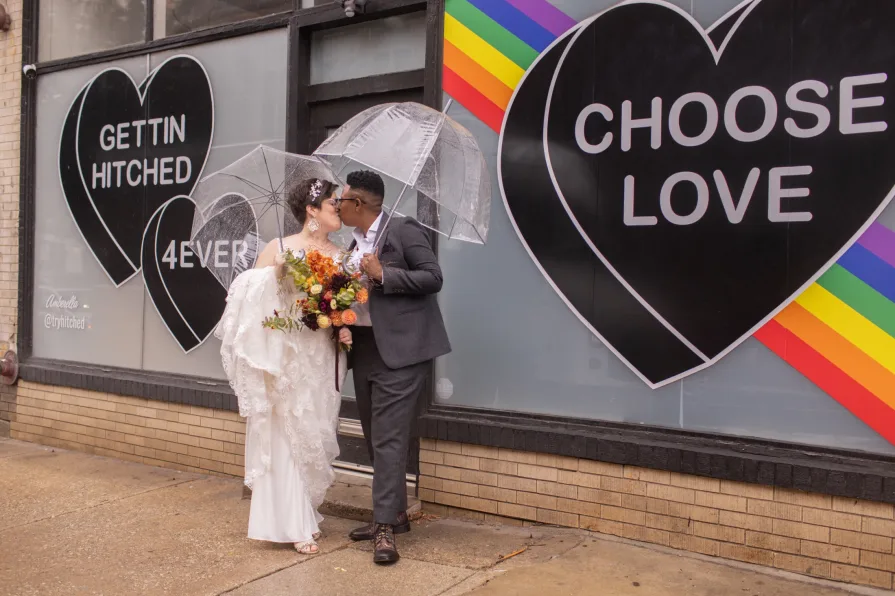 A queer, biracial couple in the rain on their wedding day in front of a Amberella XO mural.
