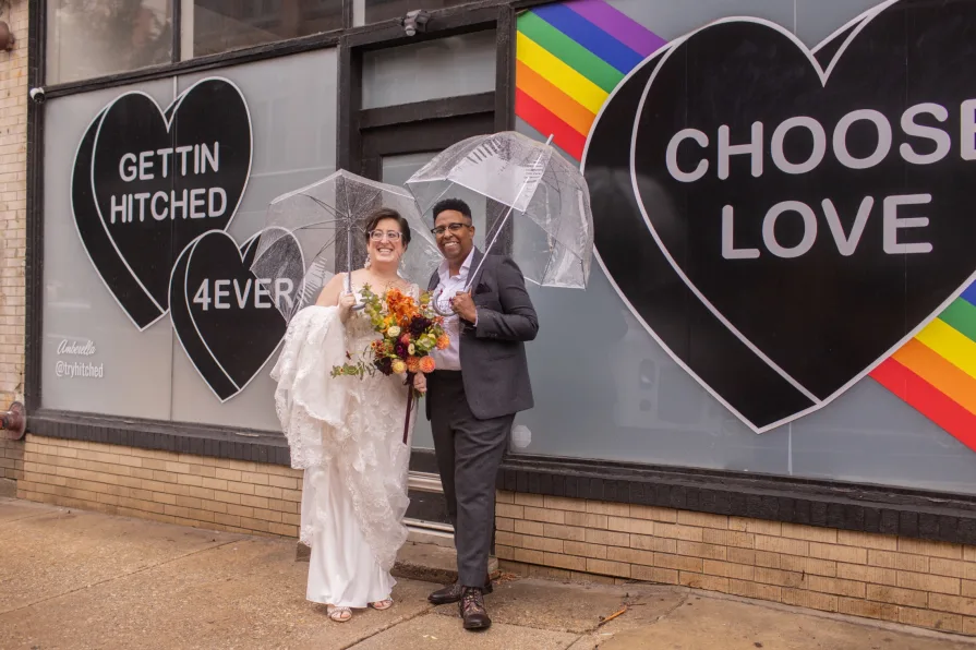 A queer, biracial couple in the rain on their wedding day in front of a Amberella XO mural.