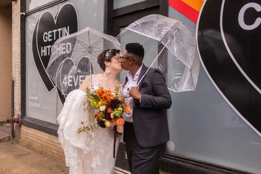 A queer, biracial couple in the rain on their wedding day in front of a Amberella XO mural.