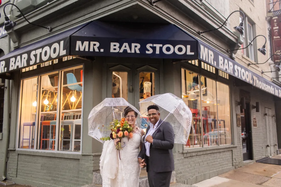 A queer, biracial couple in the rain on their wedding day in front of Mr. Bar Stool in Old City.