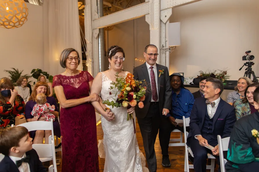 A bride walks down the aisle with her parents during an indoor ceremony at Power Plant Productions.