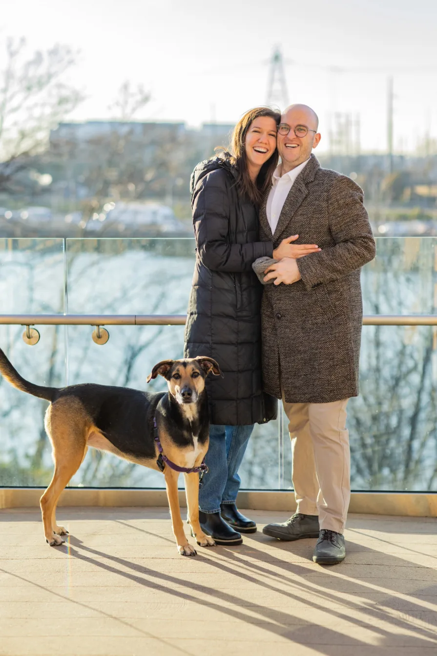 A couple smiles at the camera with their dog after their proposal.