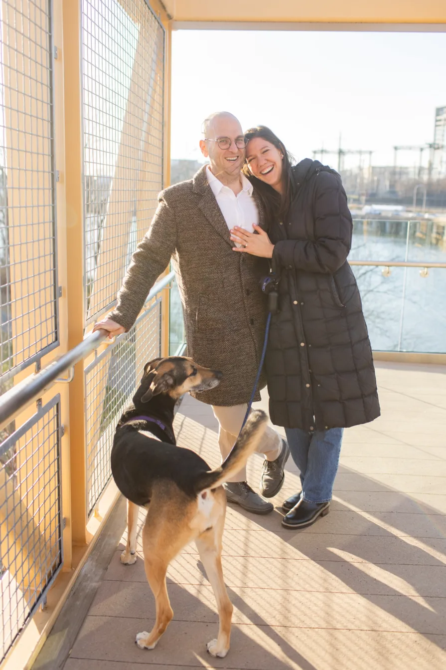 A couple smiles at the camera with their dog after their proposal.