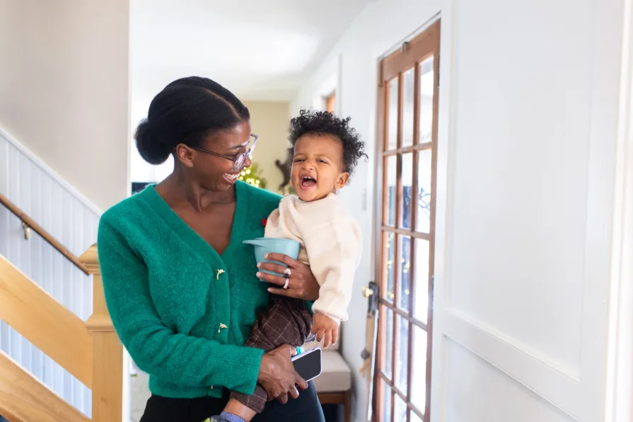 A Black mother in a green sweater smiles at her one-year-old inside their home for their family photos.