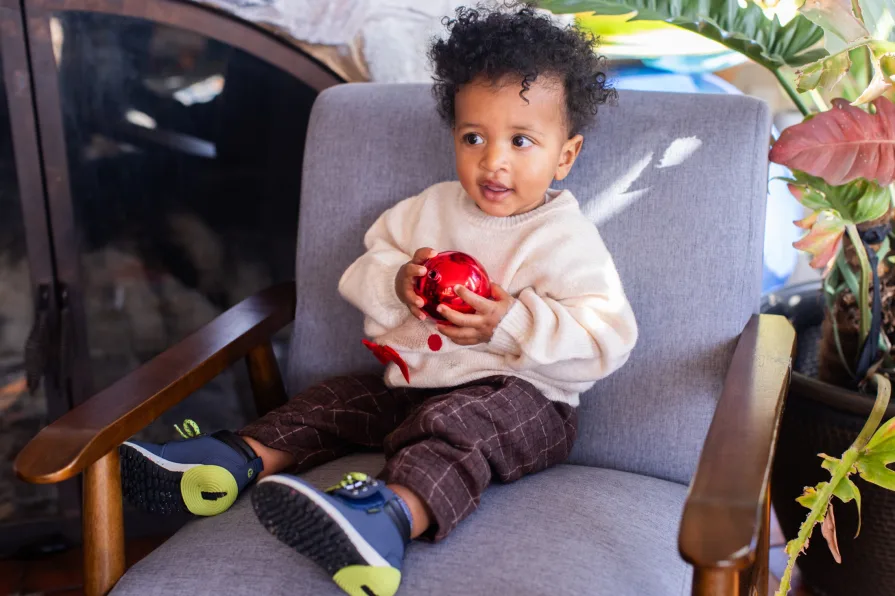 A one-year-old holds a Christmas ornament on a grey chair in his home.