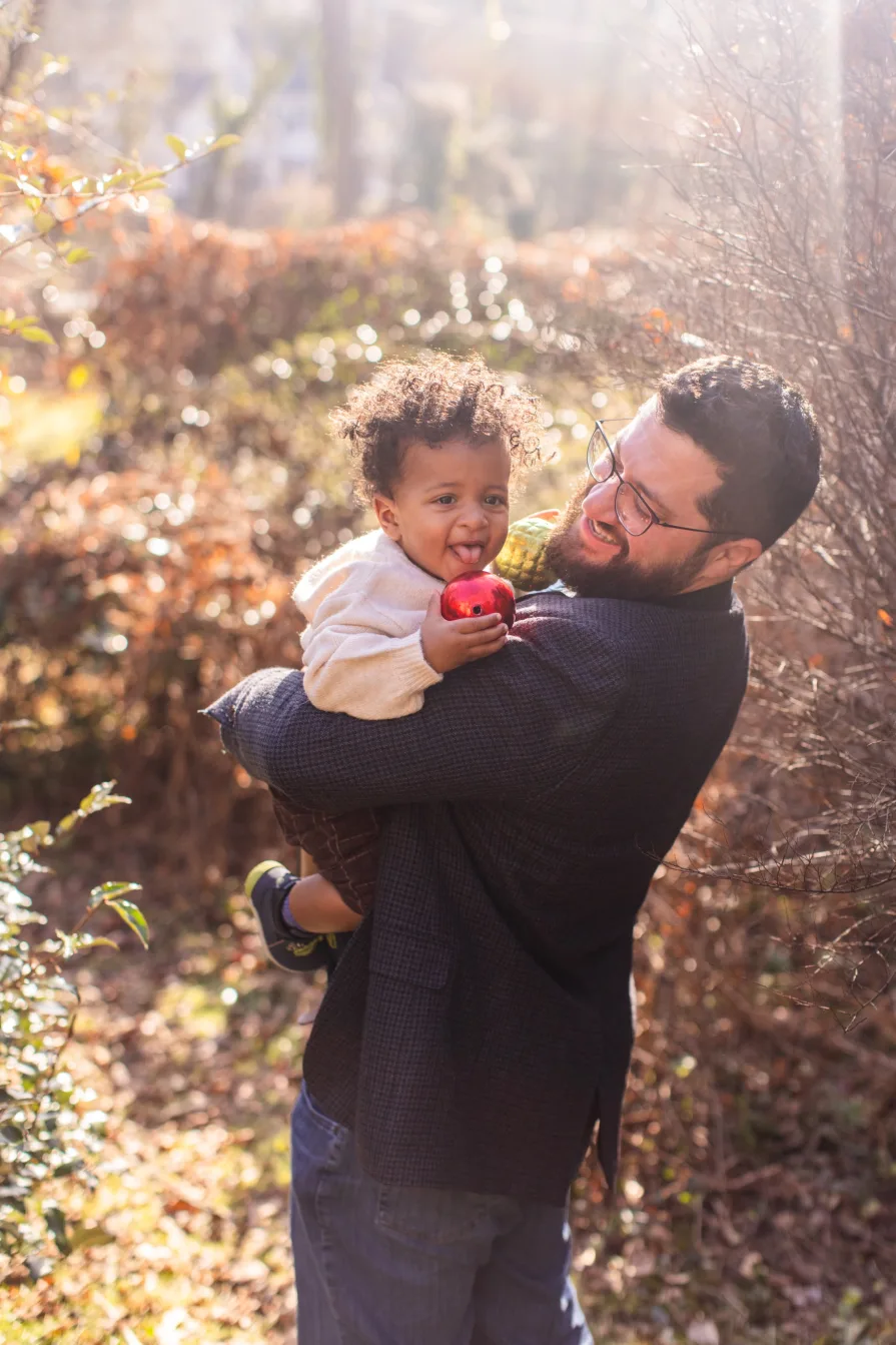 Forest being held by his dad outside while smiling in front of greens.