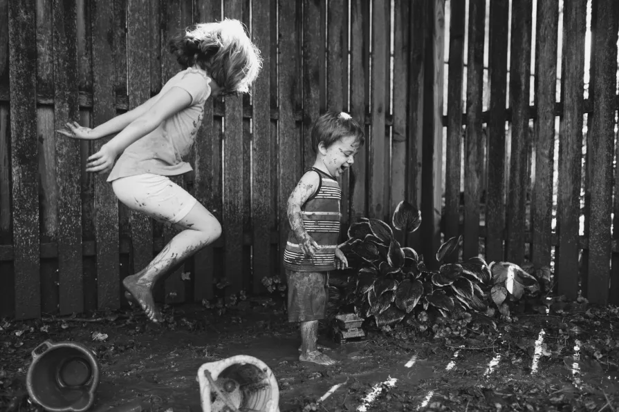 A black and white photo of two neurodivergent kids playing and jumping in the mud in their backyard.