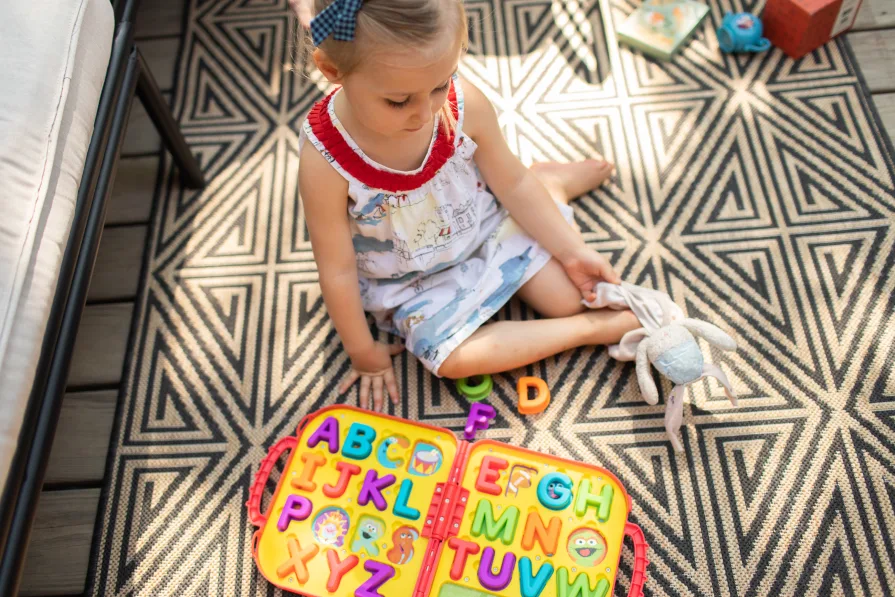 A child sits on an outdoor rug on their porch with a comfort item in their hands.