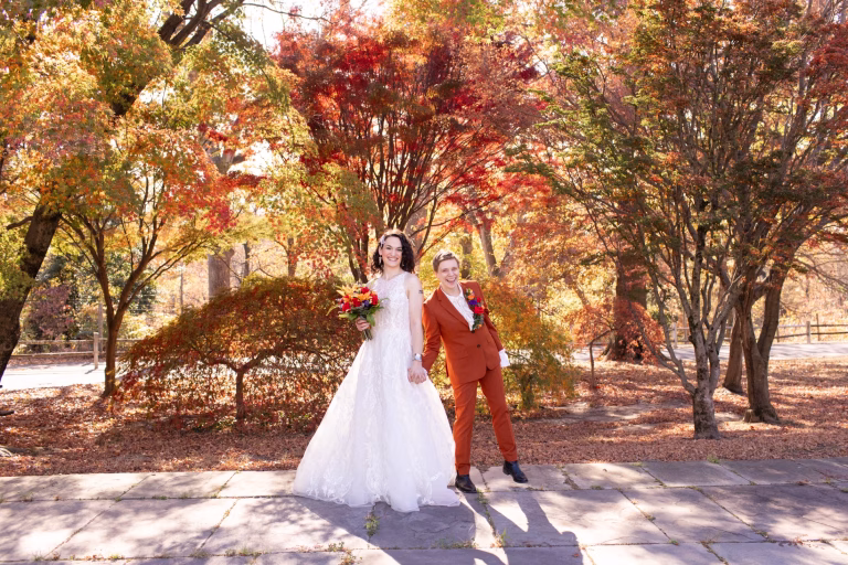 A queer couple smiling surrounded by autumnal trees before their Horticulture Center Philadelphia wedding